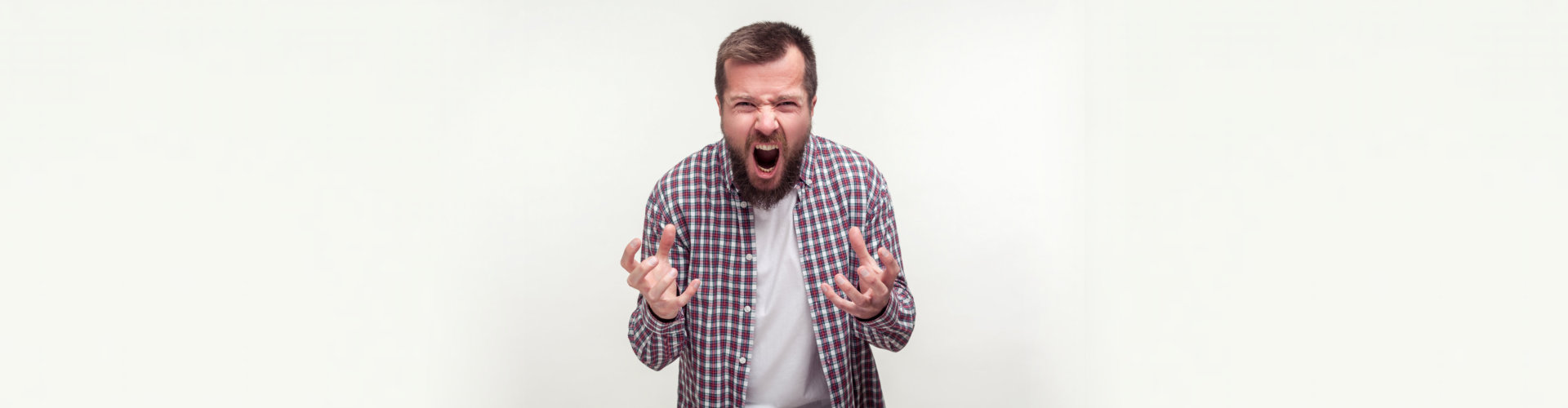 Portrait of aggressive bearded young man in plaid shirt standing with furious face full of hatred and anger, experiencing strong emotions, shouting